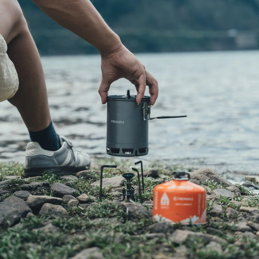 Person holding a camping stove by a lake with a propane tank nearby