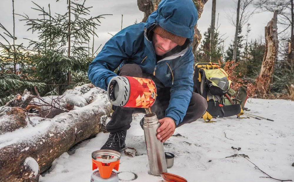 Person in a blue jacket preparing a meal with a portable stove in a snowy forest setting.
