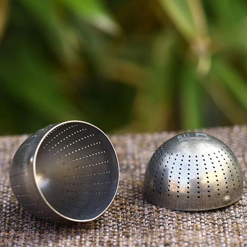 Two metallic tea infuser halves on a textured surface with a blurred green background