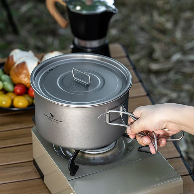 Person holding a camping pot over a portable stove with a blurred outdoor background