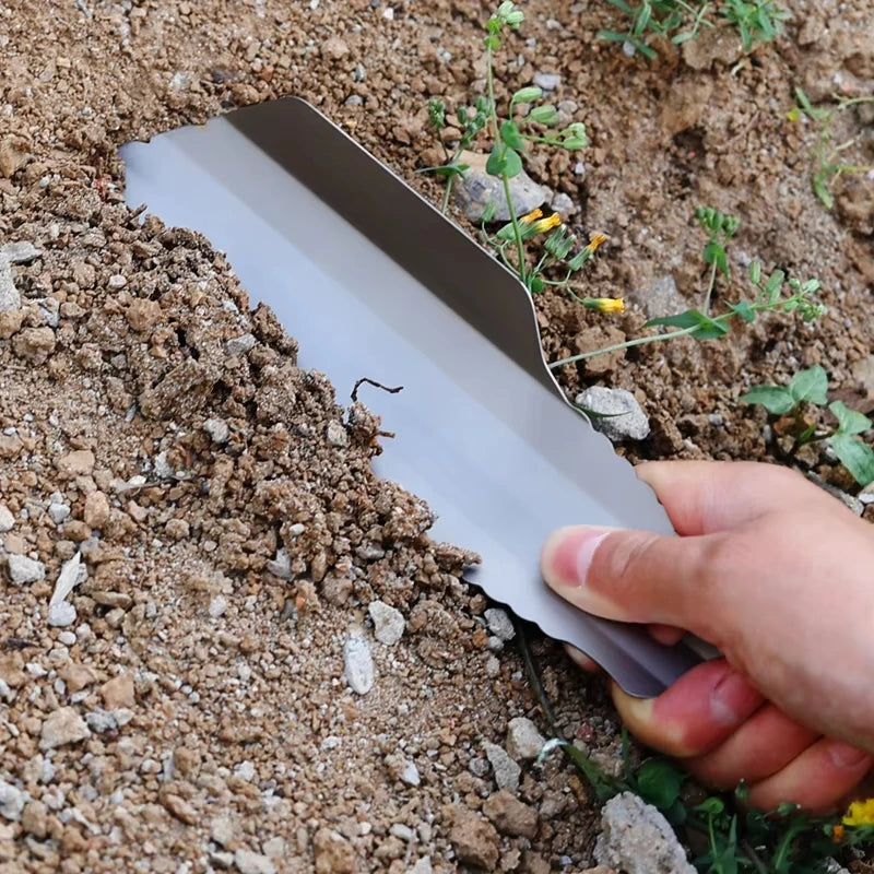 Hand using a metal tool to dig into the soil with small plants and rocks around.