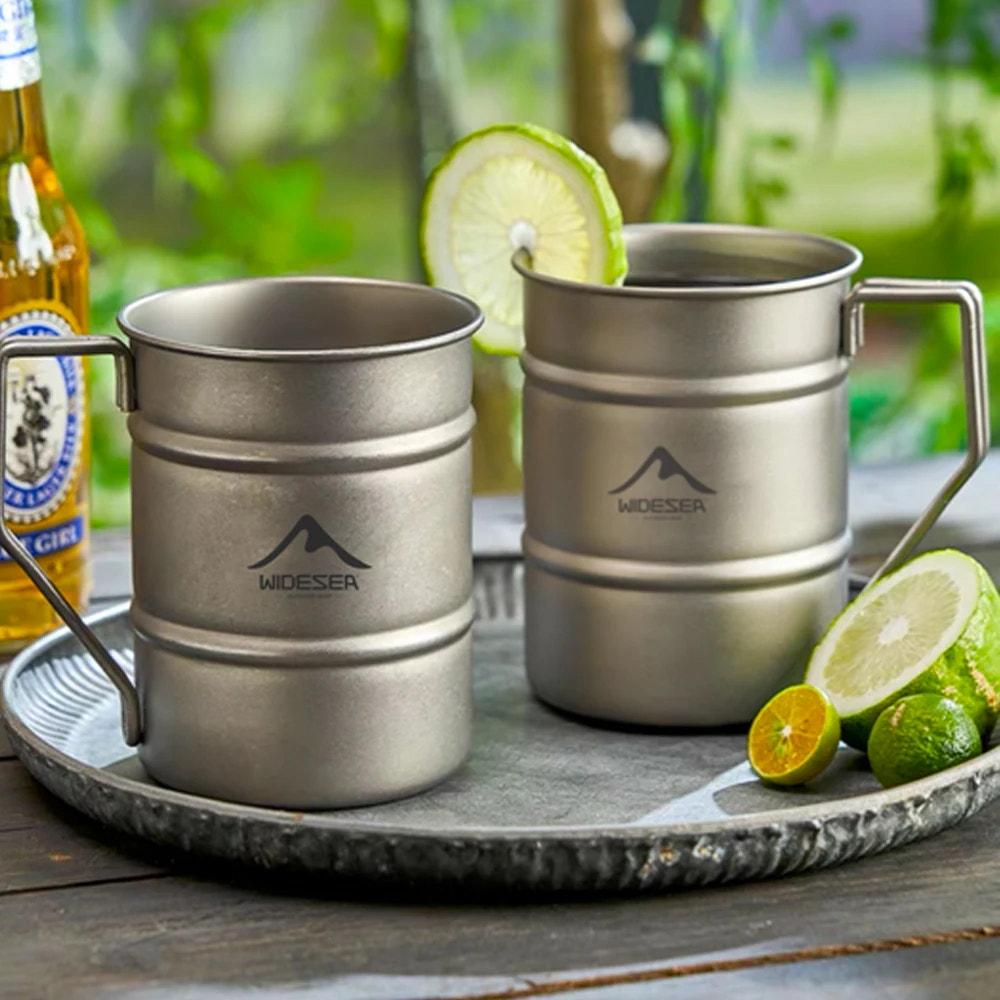 Two metal mugs with a visible brand logo on a tray with limes and a bottle in the background.