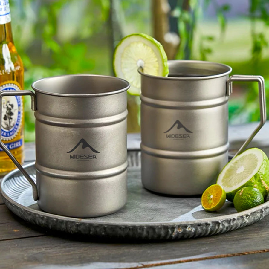 Two metal mugs with a visible brand logo on a tray with limes and a bottle in the background.
