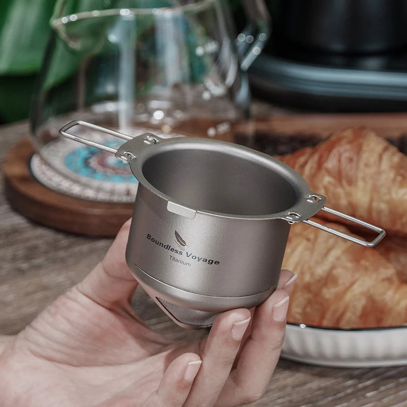 Hand holding a Titanium coffee filter with 'Boundless Voyage' branding, set against a breakfast background.