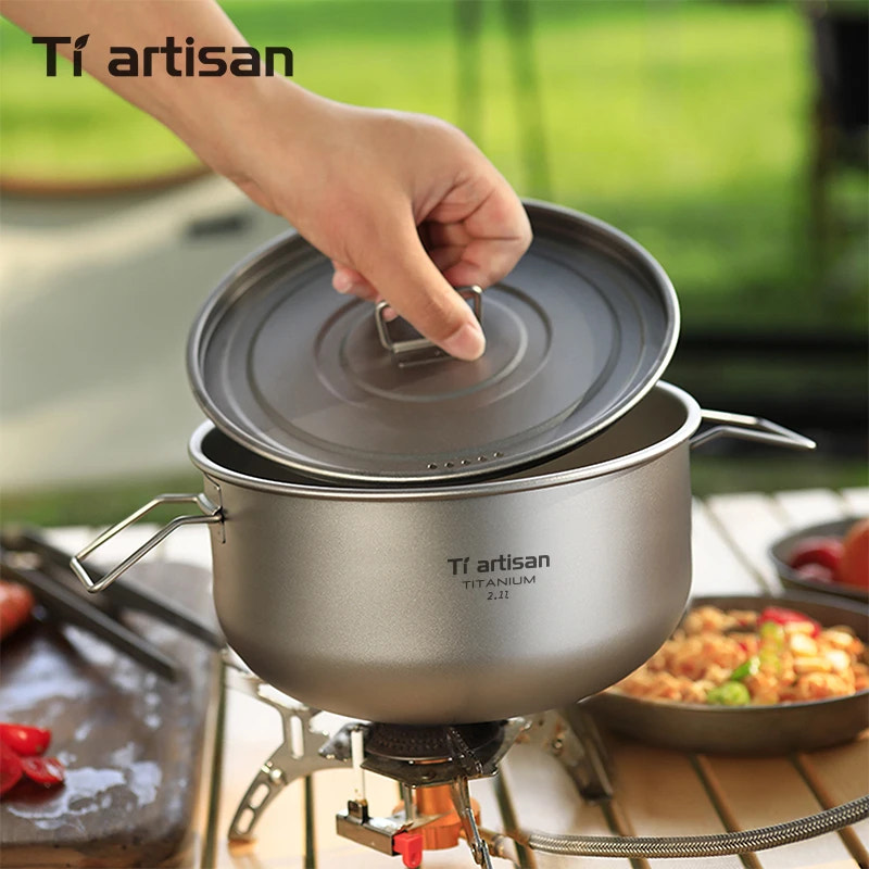 Titanium cookware set with a hand lifting a lid, set up on a camping stove outdoors.