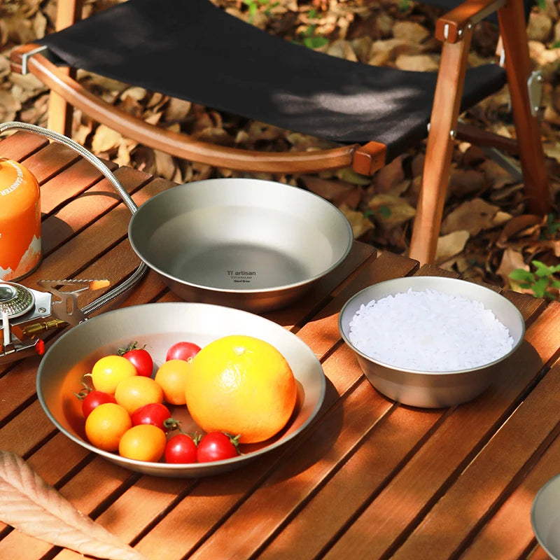 Camping cookware set on a wooden table outdoors with fruits and a chair in the background.