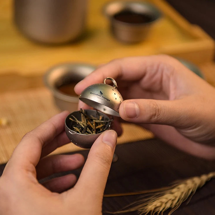 Person holding a small metal container with tea leaves on a wooden surface.