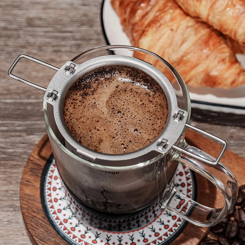 Titanium coffee filter filled with coffee on a wooden coaster with croissants in the background.