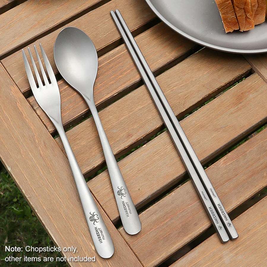 titanium spoon, fork, and chopsticks on a wooden surface with a plate of bread in the background.