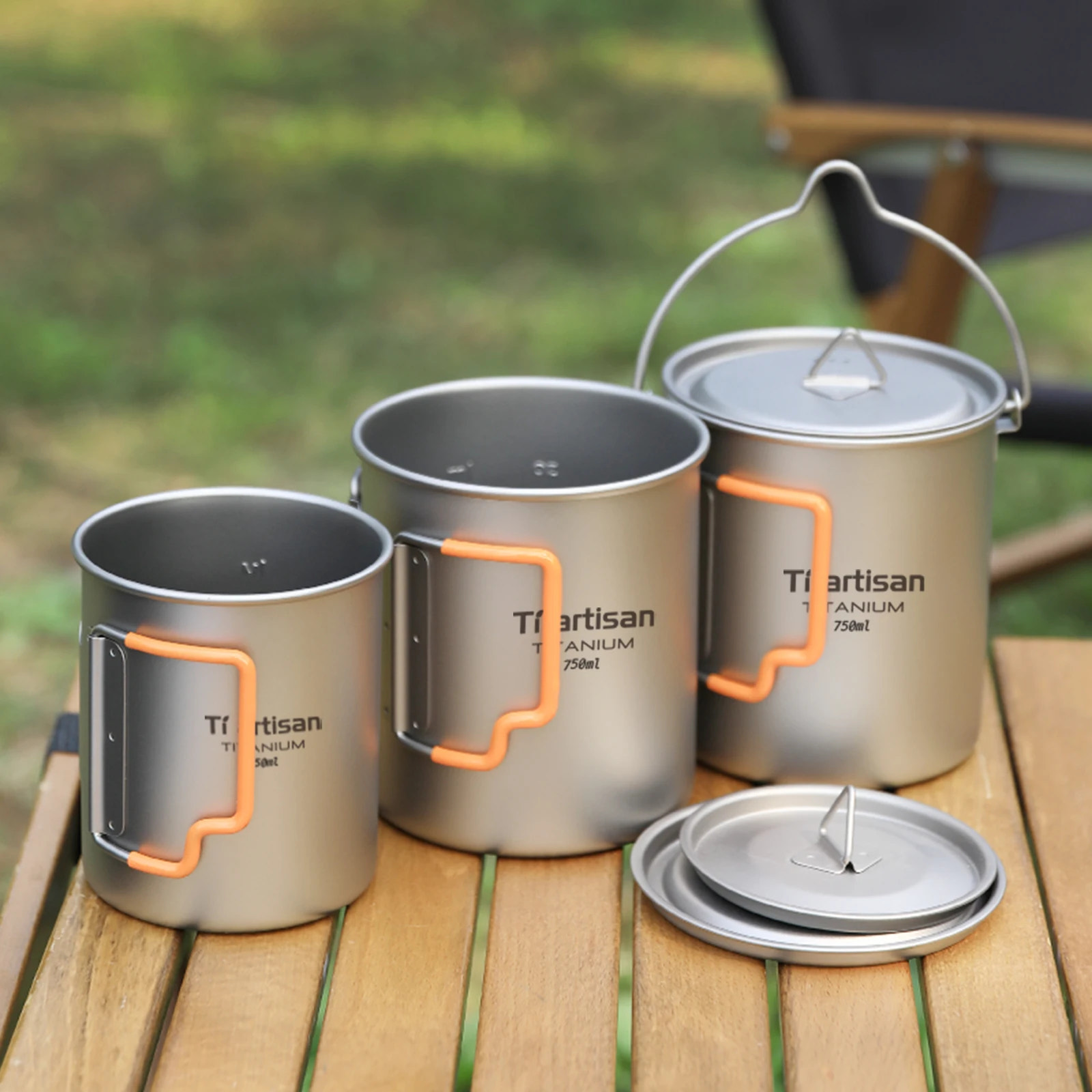 Three titanium camping mugs with orange handles on a wooden surface outdoors.