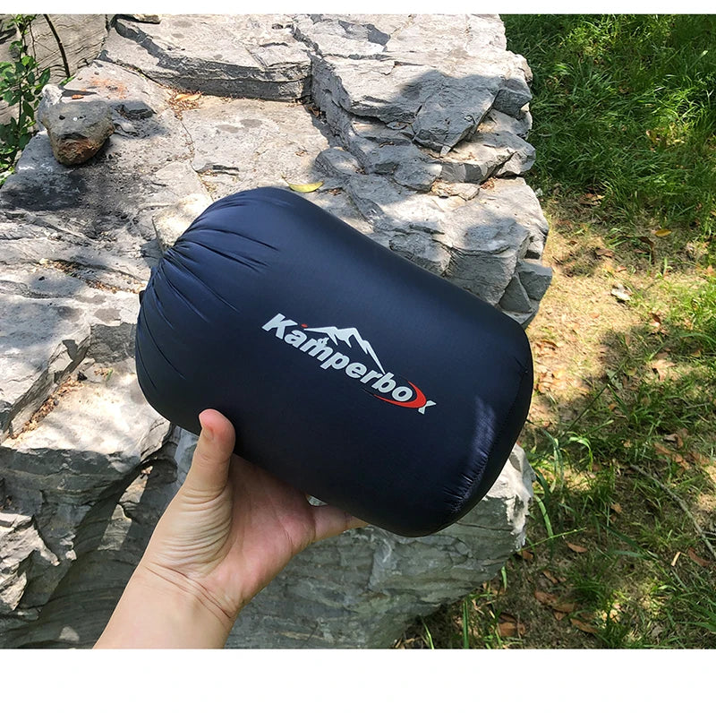 Hand holding a navy blue sleeping bag with 'Kamperbox' logo against a stone wall and grass background
