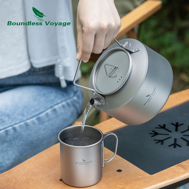 Person pouring water from a titanium camping kettle into a mug on a wooden surface with 'Boundless Voyage' branding.