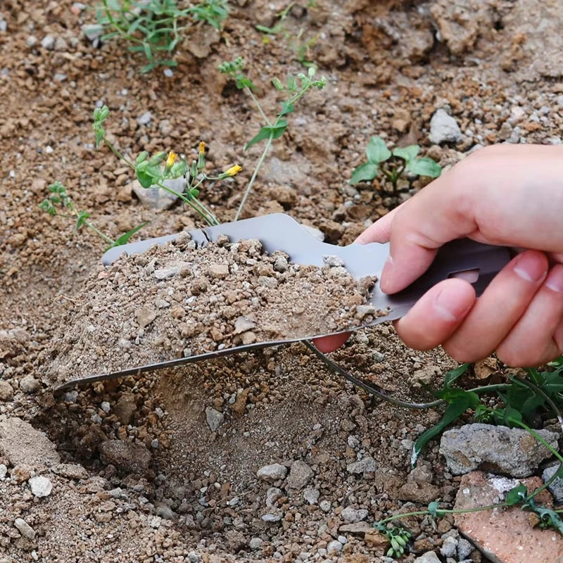 Hand using a small shovel to dig into the ground with some plants around