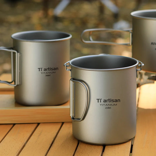 Titanium mugs with handles on a wooden surface, featuring 'Ti artisan' branding.