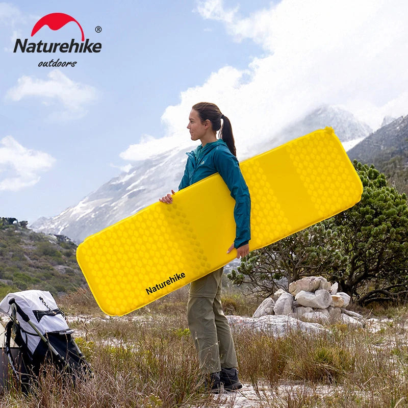 Person holding a yellow inflatable mattress with Naturehike branding in a mountainous outdoor setting.