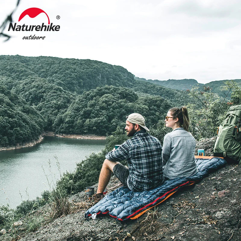 Two people sitting on a Naturehike quilt overlooking a scenic landscape with mountains and water.