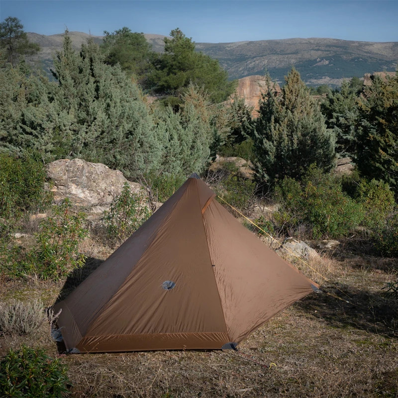 Brown tent set up in a forested area with trees and mountains in the background