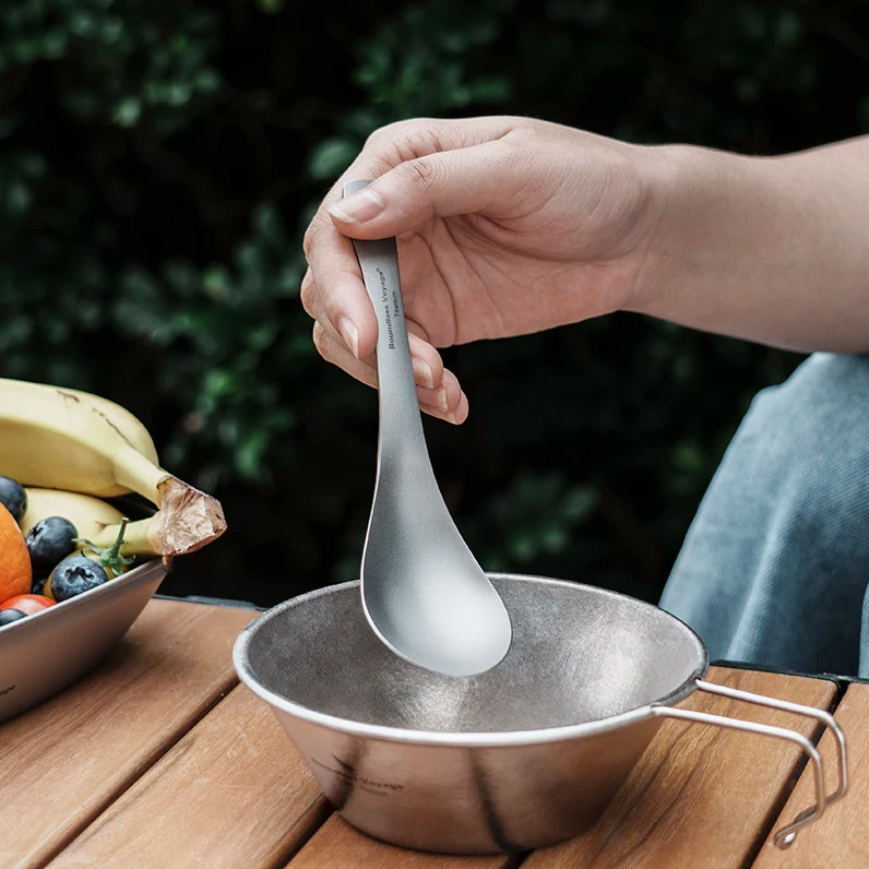 Person holding a titanium spoon over a metal bowl with a blurred outdoor background