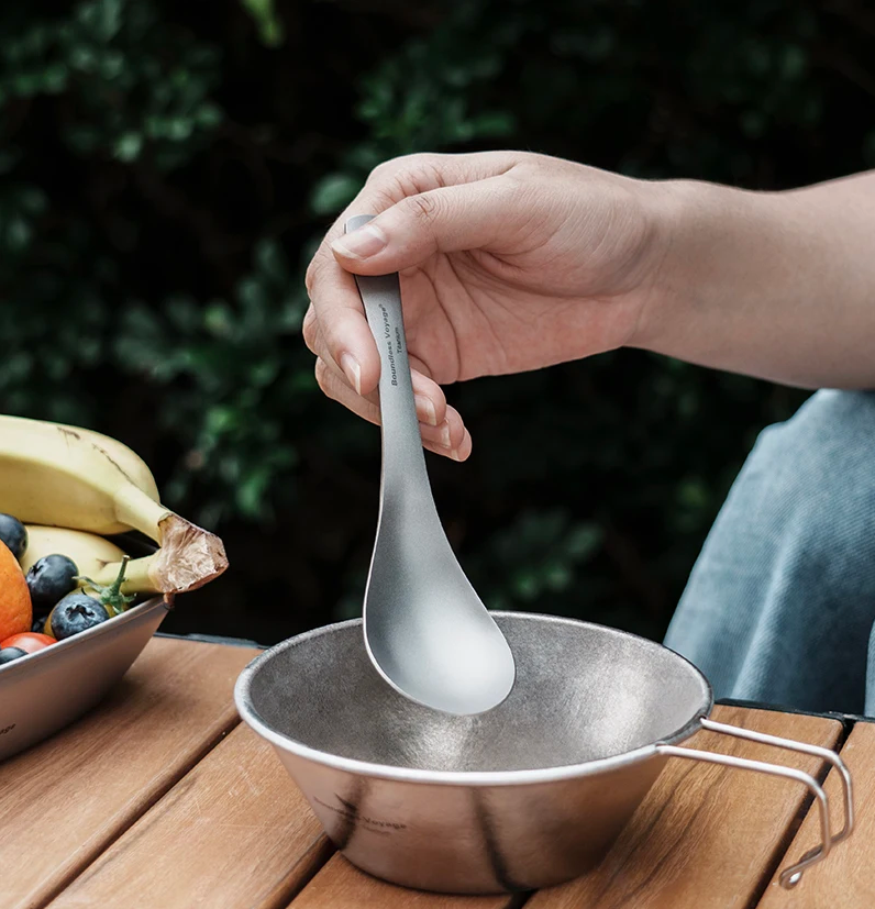 Person holding a titanium spoon over a metal bowl with a blurred outdoor background