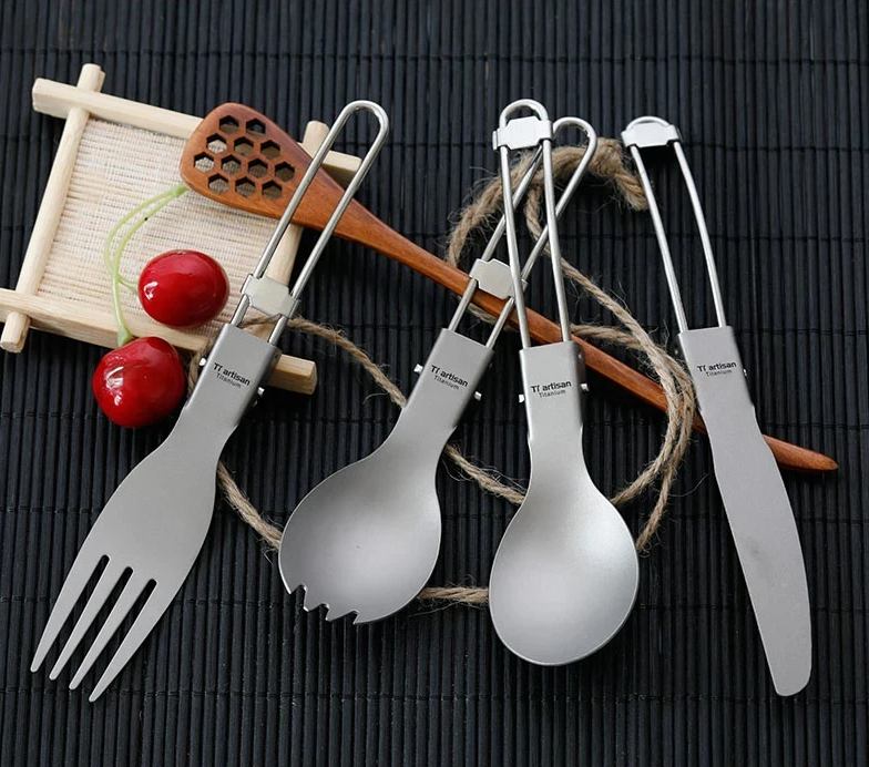 Set of titanium cutlery including fork, spoon, and knife on a dark surface with wooden utensils and cherries.