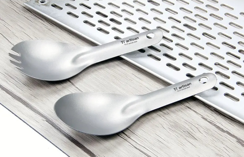 Two titanium spoons with 'Ti artisan' branding on a wooden surface.