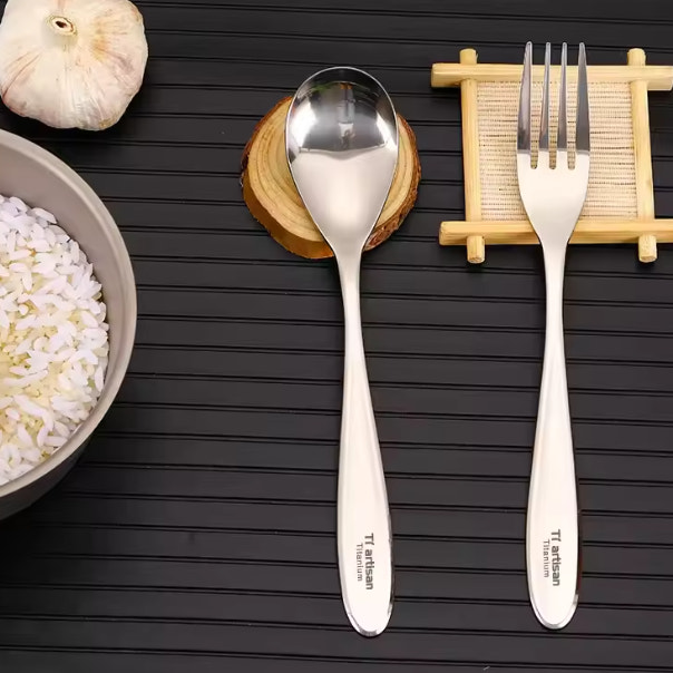 Titanium spoon and fork on a dark wooden surface with a bowl of rice and garlic.