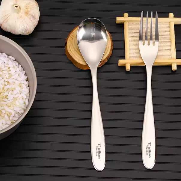 Titanium spoon and fork on a dark wooden surface with a bowl of rice and garlic.