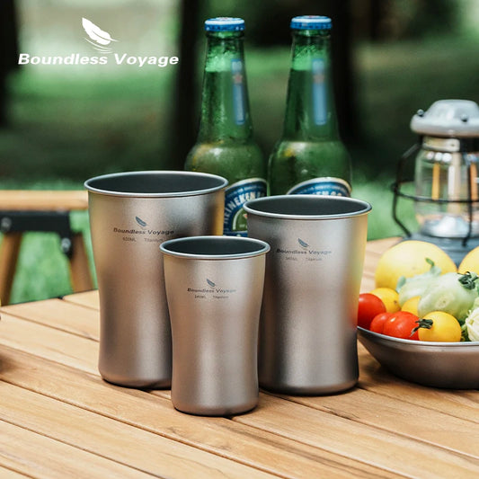 Three titanium cups on a wooden table with Boundless Voyage branding, surrounded by bottles and fruits.