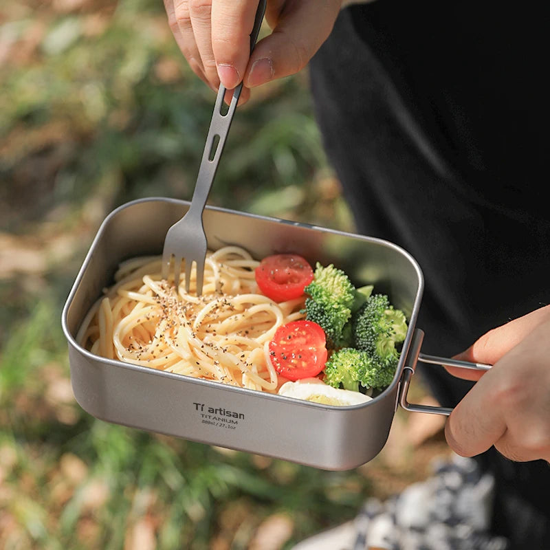 Person holding a portable meal with spaghetti, vegetables, and a fork outdoors.