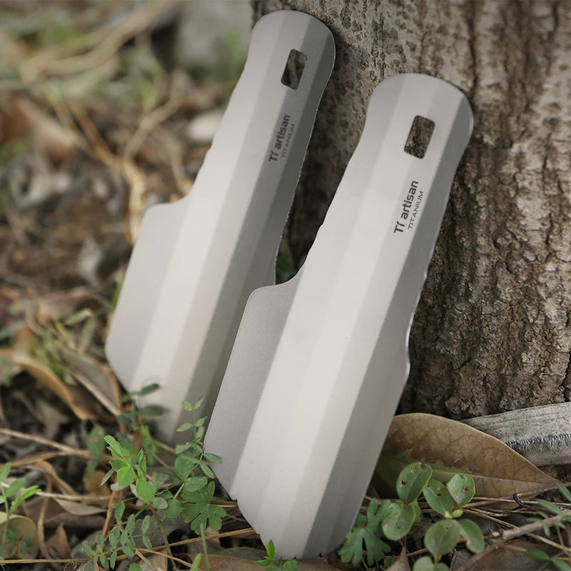 Two titanium tools leaning against a tree trunk with grass and leaves in the foreground.