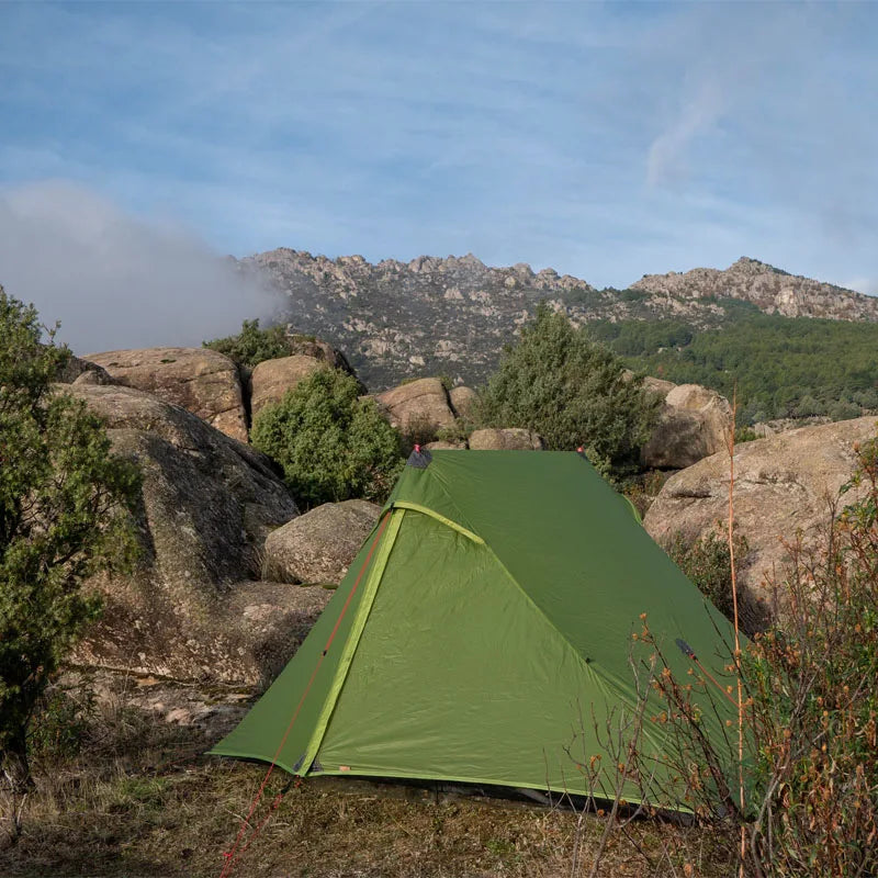 Green tent set up in a rocky, mountainous area with trees and a clear sky.