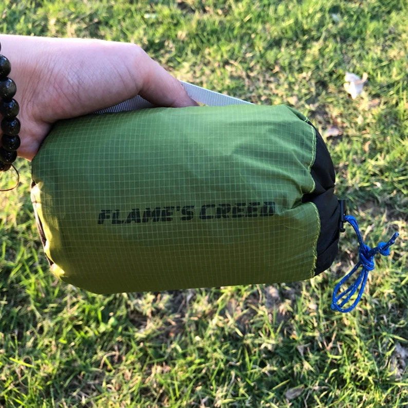 Green stuff sac with 'Flame's Creed' branding held by a person on grass.