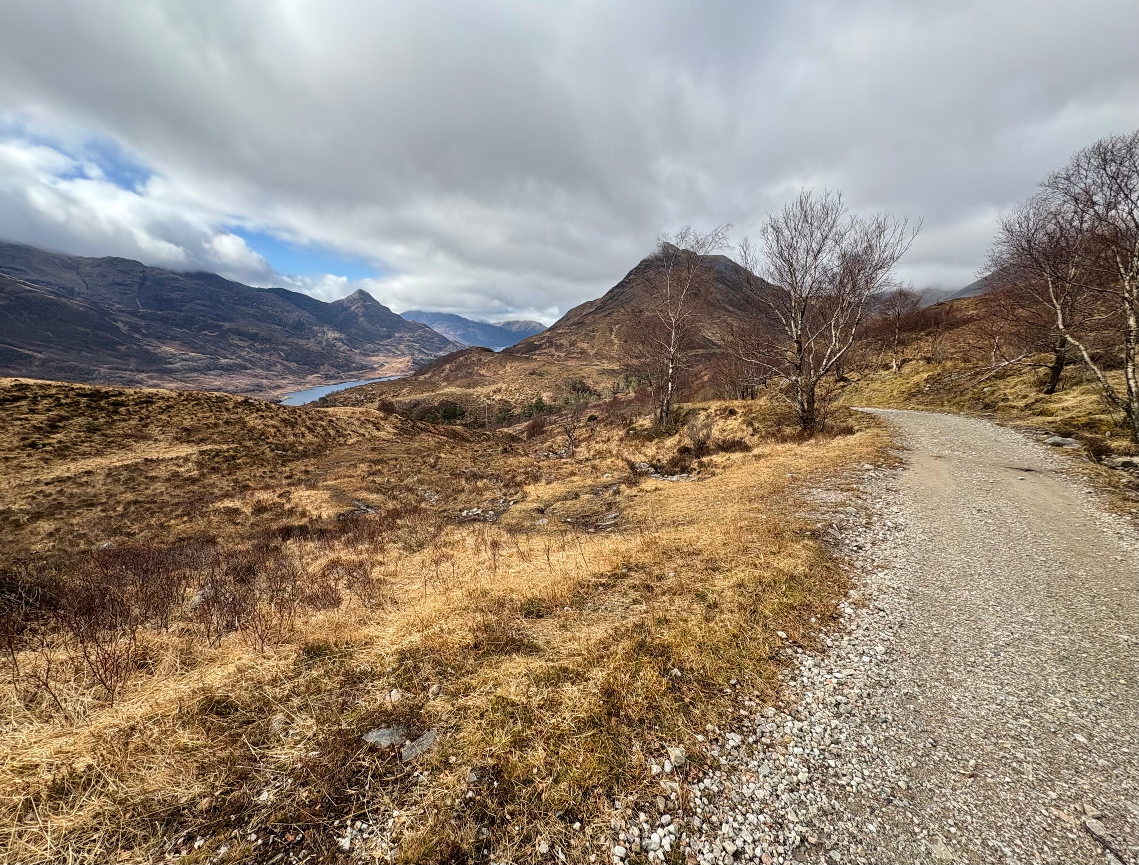 Winding dirt road through a mountainous landscape with bare trees and cloudy sky.