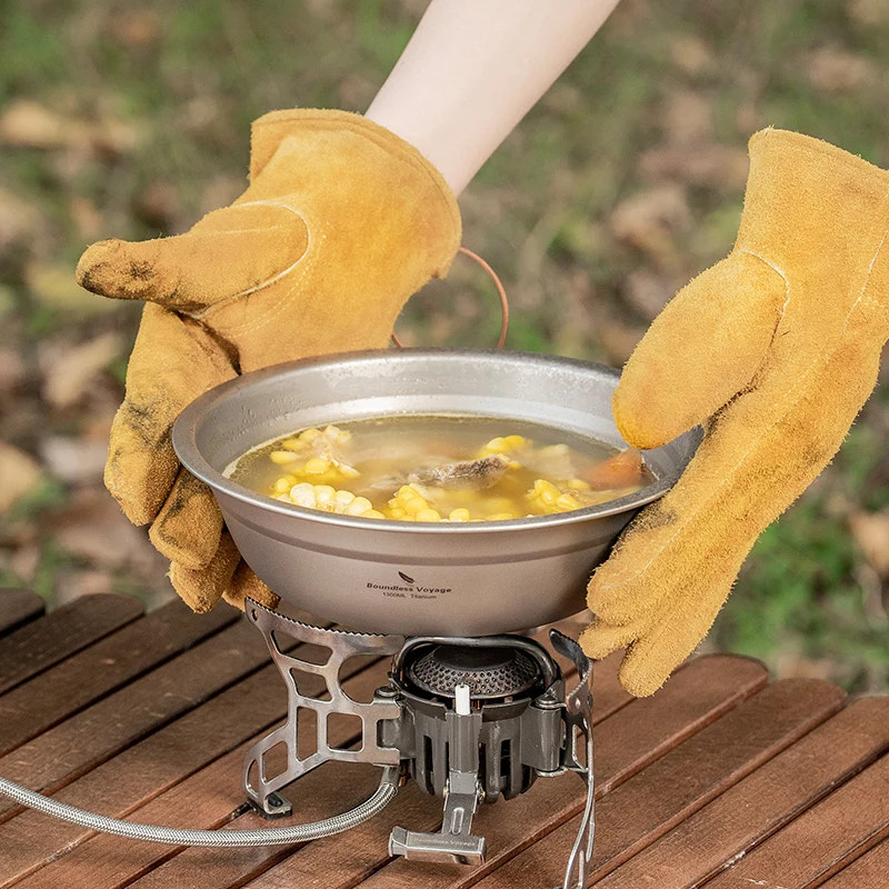 Person wearing yellow gloves holding a bowl of soup over a portable stove on a wooden surface.