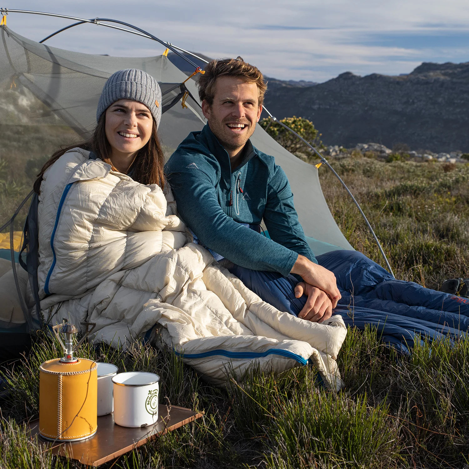Two people sitting under a tent in a mountainous area with camping gear.