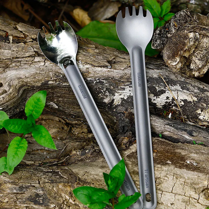 Two titanium Spork utensils on a wooden surface with green leaves.