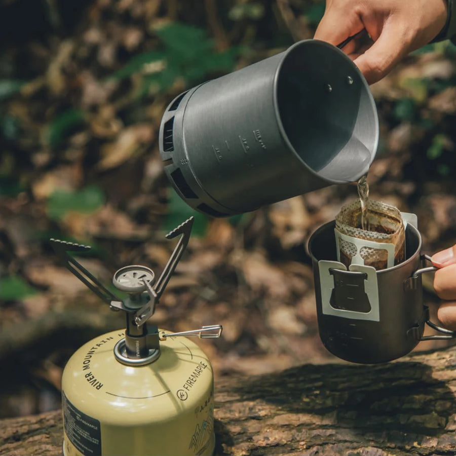 Person pouring coffee from a camping pot into a filter over a gas stove in a forest setting.