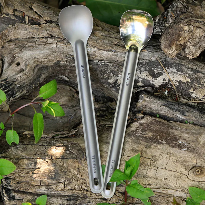 Two titanium spoons on a wooden surface with green leaves.