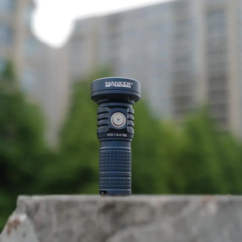 Blue flashlight with 'MANKER' branding on a stone surface with blurred greenery and building in the background