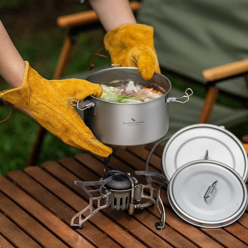 Person wearing yellow gloves holding a pot over a portable stove with camping cookware on a wooden surface.