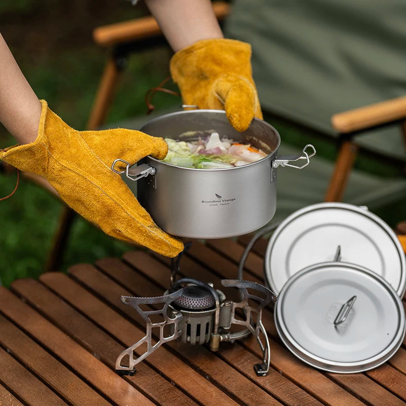 Person wearing yellow gloves holding a pot over a portable stove with camping cookware on a wooden surface.