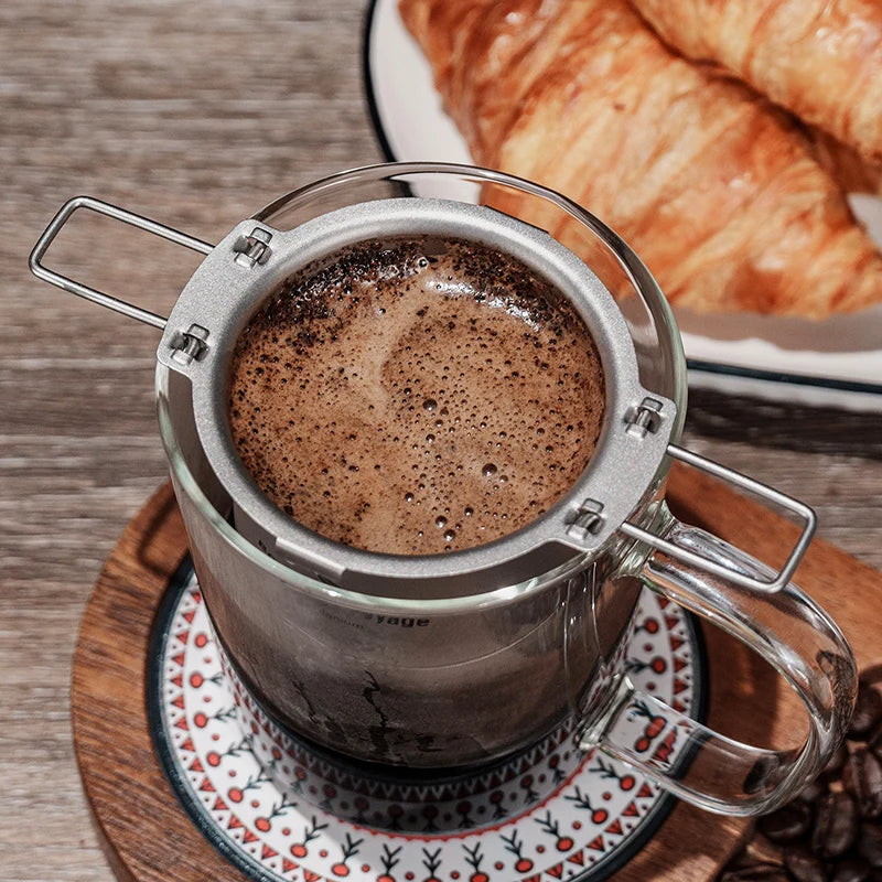 Titanium coffee filter filled with coffee on a wooden coaster with croissants in the background.