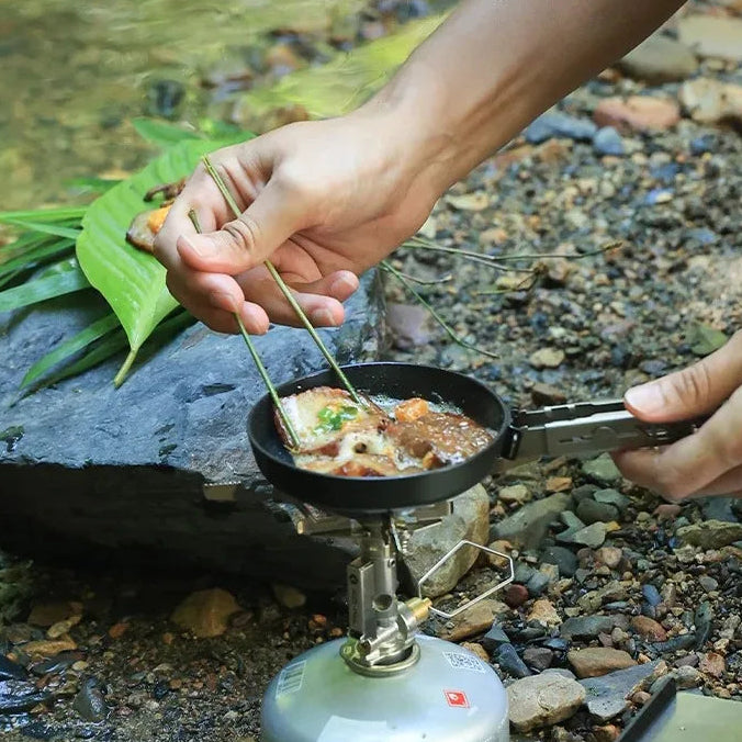 Person cooking outdoors using a portable stove and frying pan on a natural background.