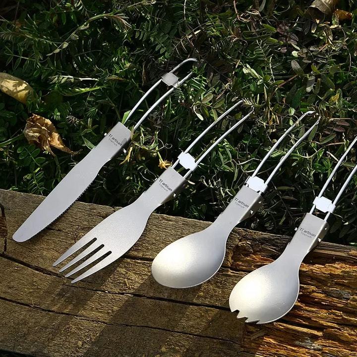 Set of titanium camping utensils including a knife, fork, and two spoons on a wooden surface with greenery in the background.