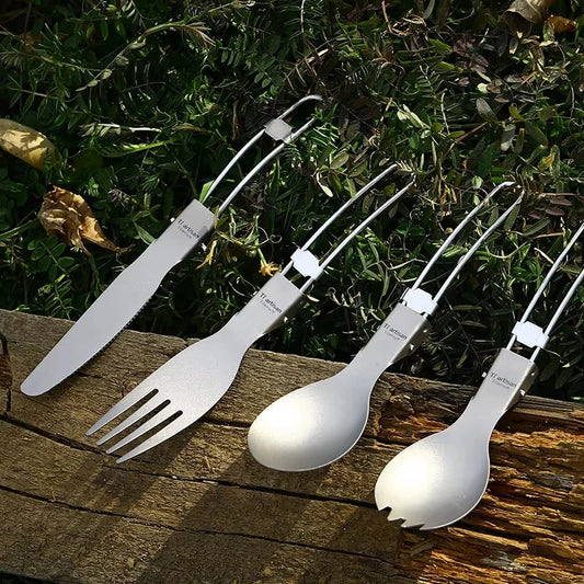 Set of titanium camping utensils including a knife, fork, and two spoons on a wooden surface with greenery in the background.