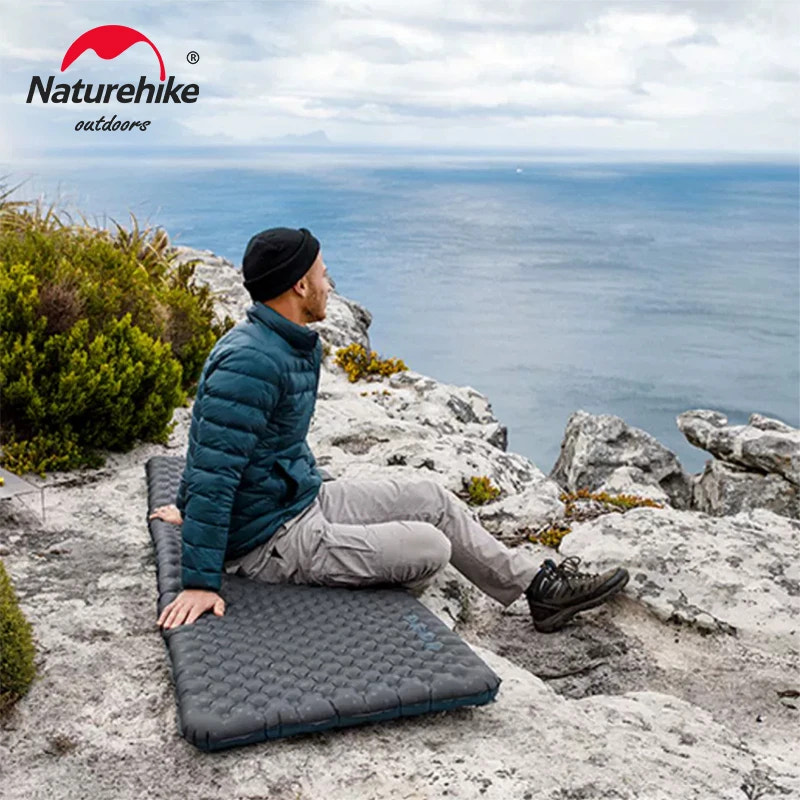 Person sitting on a Naturehike outdoor mat by the ocean with the Naturehike logo visible.