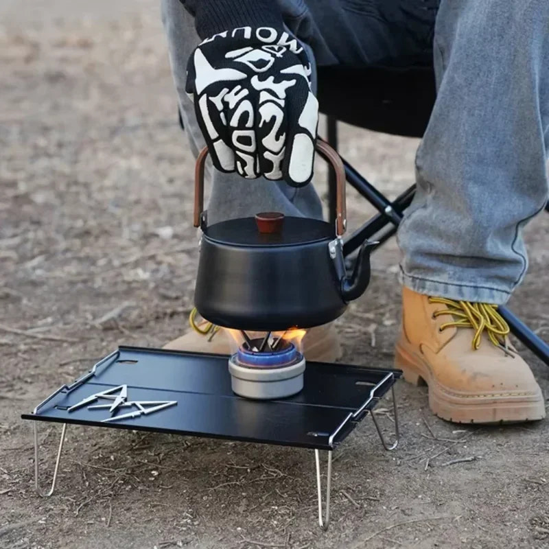 Person using a portable camping stove with a kettle on a camping site.