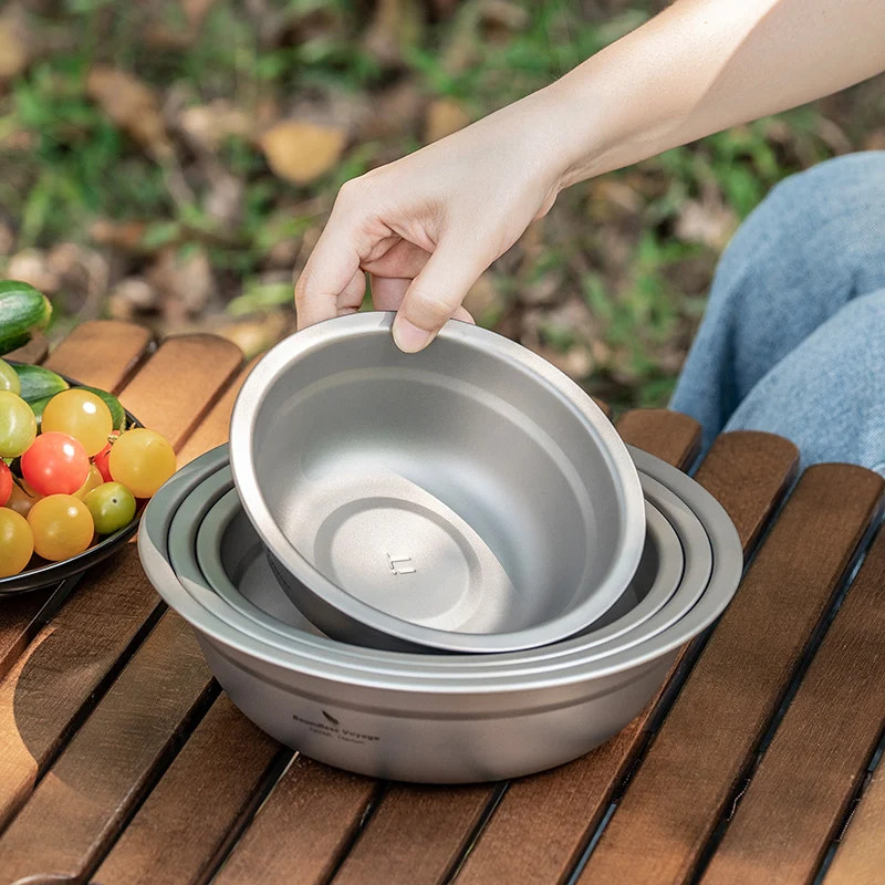 Stack of titanium bowls on a wooden table with a hand reaching for one, outdoors.