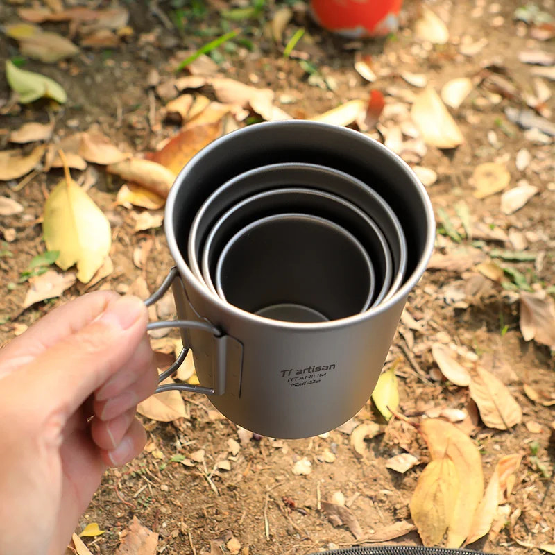 Set of metal measuring cups held by a hand outdoors on a leafy ground.