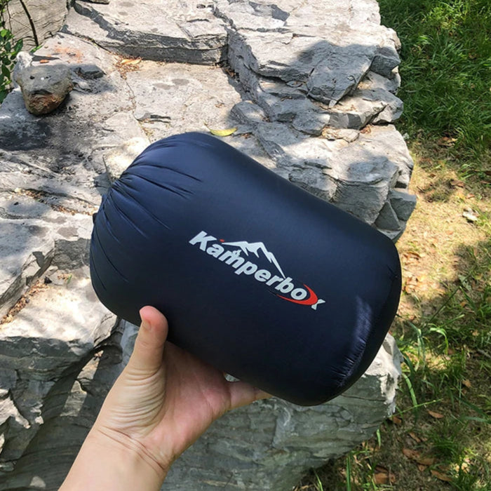 Hand holding a navy blue sleeping bag with 'Kamperbox' logo against a stone wall and grass background